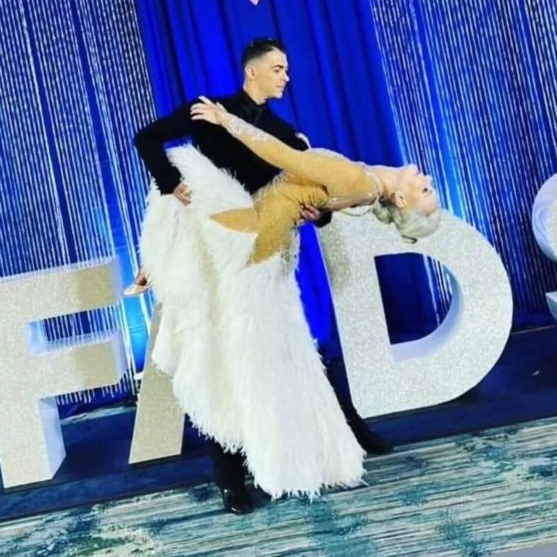 Tatiana Vaccaro in a white feathered gown held in a deep, sweeping backbend by her partner against a blue stage backdrop