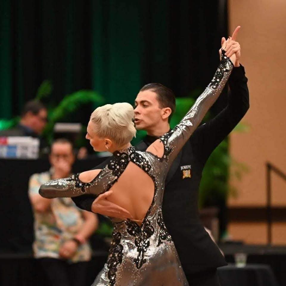 Tatiana Vaccaro in a silver ballroom gown, held in a dramatic closed frame with her partner on the competition floor