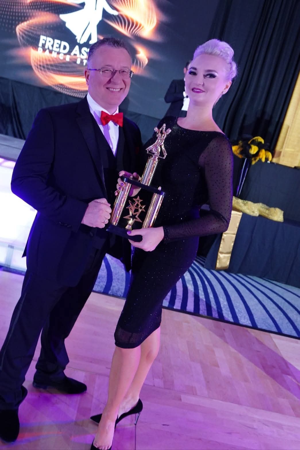 Tatiana Vaccaro in an elegant black dress holding a Fred Astaire competition trophy beside a presenter