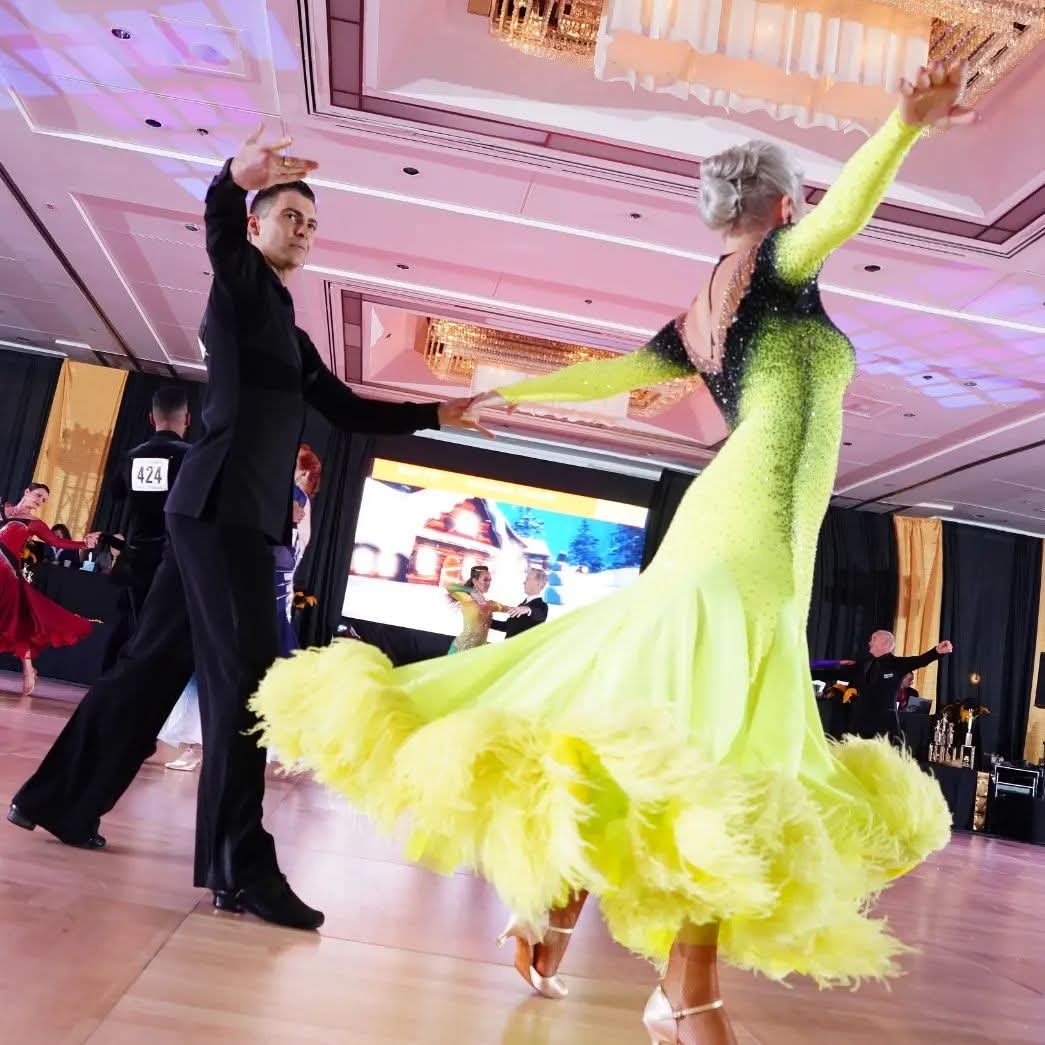Tatiana Vaccaro in a yellow feathered ballroom gown, mid-turn with her partner on the competition floor