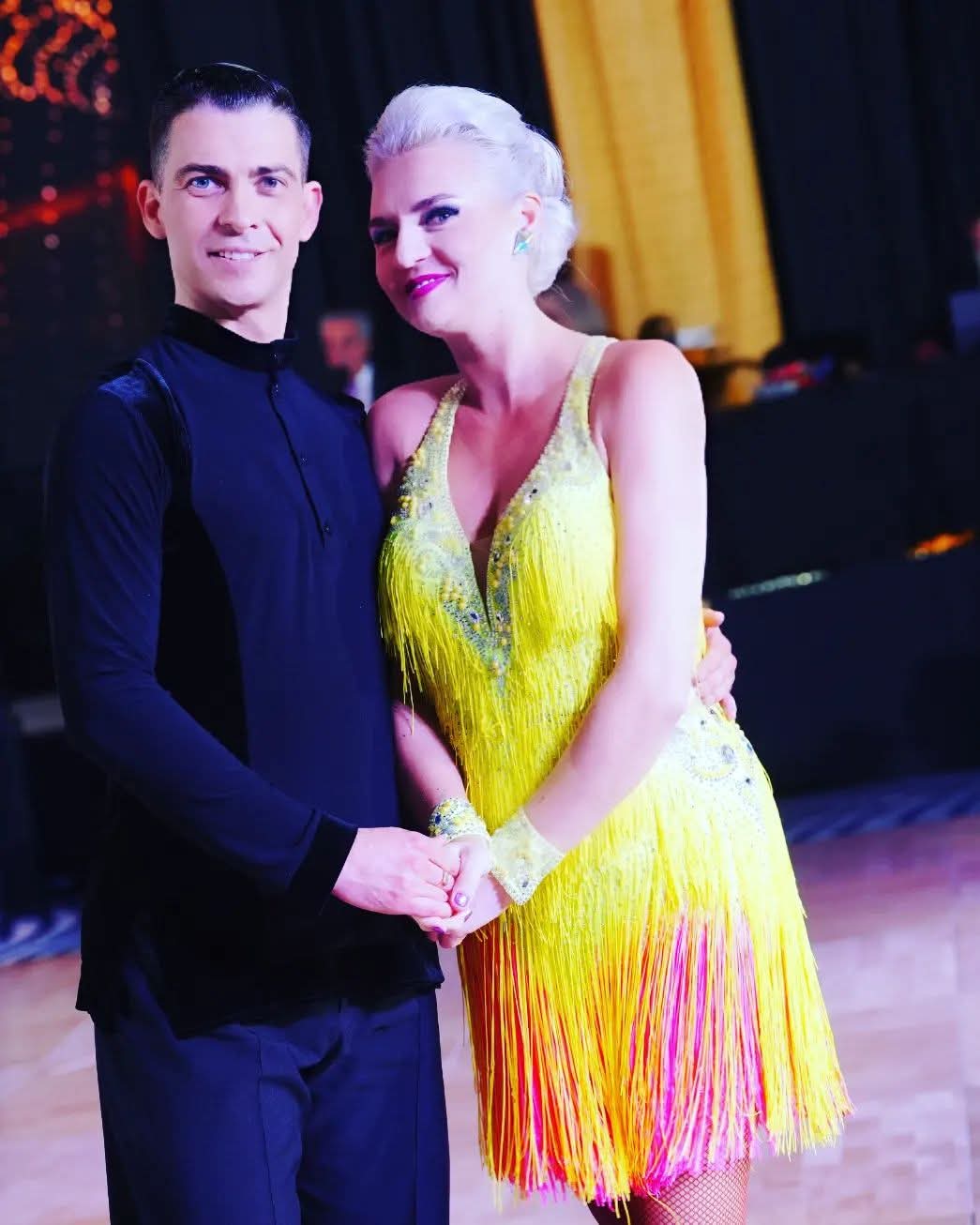 Tatiana Vaccaro in a yellow fringe Latin dress holding hands with her partner at a ballroom competition