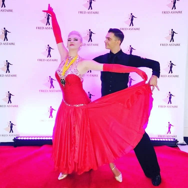 Tatiana Vaccaro in a flowing red ballroom gown, wearing a medal and posed with her partner at a Fred Astaire event